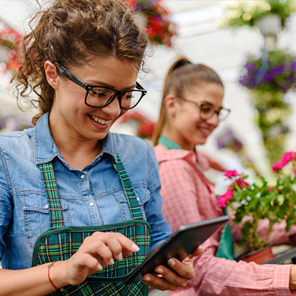 Zwei Frauen lachen. Eine betrachtet ein Tablet, während die andere einen Blumentopf mit Pflanze weg räumt - entspanntes Smart Gardening mit Kaisers Gartenbau